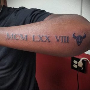Фото татуировки на руке с римскими цифрами 'MCM LXX VIII' и изображением головы быка. Черный рисунок выглядит брутально и стильно.