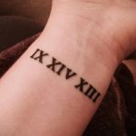 Фото татуировки с римскими цифрами 'IX XIV XIII' на запястье. Черный рисунок выглядит четко и минималистично на светлой коже.
