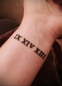 Фото татуировки с римскими цифрами 'IX XIV XIII' на запястье. Черный рисунок выглядит четко и минималистично на светлой коже.