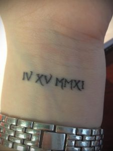Фото татуировки с римскими цифрами IV XV MM XXI на запястье. Минималистичный дизайн, выполненный черными чернилами.