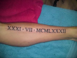 Фото тату: на предплечье выгравированы римские цифры 'XXXI VII MCLXXXII' черными чернилами. Минималистичный стиль, крупная татуировка.