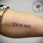 Фото тату: римские цифры XX.III.MM на плече, выполненные черными чернилами, с логотипом тату-студии.