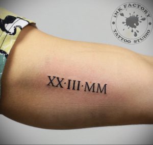 Фото тату: римские цифры XX.III.MM на плече, выполненные черными чернилами, с логотипом тату-студии.