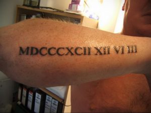 Фото тату: римские цифры MDCCCXCII XII VI III на предплечье мужчины, выполненные черными чернилами, классический стиль.