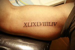 Фото тату: черные римские цифры XLIX.LVII.ILIV на бицепсе мужчины, выполненные четкими линиями.