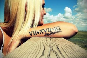 Фото тату: римские цифры VII.XVII.XI и подпись на руке девушки на фоне голубого неба и поля.