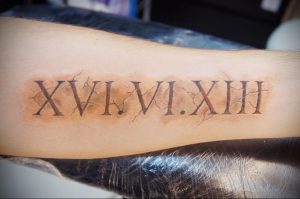 Фото тату: римские цифры XVI.VI.XIII на предплечье, выполненные коричневыми чернилами, с эффектом потертости и небольшими трещинами.