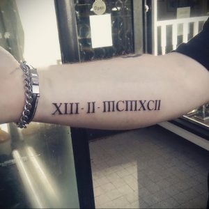 Фото тату: черные римские цифры 'XIII-II-MCMXII' на предплечье. Классический и минималистичный дизайн, подчеркивающий индивидуальность. Четкие линии и аккуратное исполнение.
