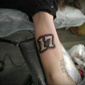 Фото тату на предплечье: число 17, выполненное в готическом стиле с использованием черного цвета и теней. Выразительный рисунок татуировки.