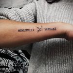 Фото тату на предплечье: черные римские цифры MCMXLVIII и MCMXXX с изображением летящей птицы между ними. Минималистичный рисунок татуировки.