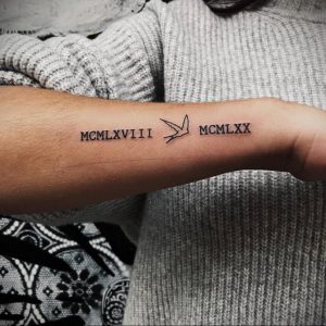 Фото тату на предплечье: черные римские цифры MCMXLVIII и MCMXXX с изображением летящей птицы между ними. Минималистичный рисунок татуировки.