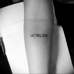 Черно-белое фото тату с римскими цифрами MCMXLIV на предплечье. Простой, элегантный дизайн, выполненный четкими линиями.