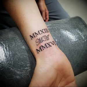 Фото татуировки с римскими цифрами MMXIII, инициалами M&A и MMXVI на запястье. Черно-белый рисунок татуировки, выполненный четкими линиями.