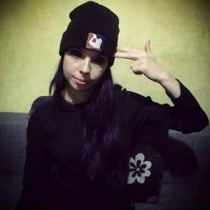 Фотография девушки в черной шапке и футболке, показывающей жест "пистолет". Выражение лица дерзкое, фиолетовые пряди волос.
