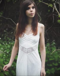 Фотография девушки в белом платье, стоящей на фоне зеленого леса. Нежный и романтичный образ, подчеркнутый естественным освещением.