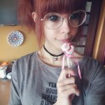 Фотография девушки с рыжими волосами в очках и с леденцом в руке. Девушка смотрит в камеру с легкой улыбкой.