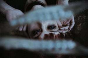 Фотография девушки с рыжими волосами, лежащей на земле и смотрящей в камеру сквозь решетку. Ее глаза выражают грусть и тоску.