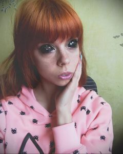 Фотография девушки с рыжими волосами в розовой толстовке с кошачьим принтом. Девушка смотрит в камеру с легкой улыбкой.