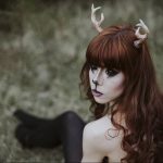 Фотография девушки с рыжими волосами и рогами, украшенными цветами. На лице девушки нанесен макияж, имитирующий мордочку животного.