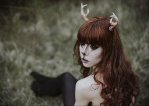 Фотография девушки с рыжими волосами и рогами, украшенными цветами. На лице девушки нанесен макияж, имитирующий мордочку животного.