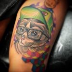 Фото тату кота в очках и кепке на плече. Яркий рисунок в стиле old school с использованием насыщенных цветов и четких контуров. Забавный и оригинальный дизайн.