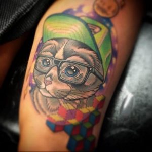 Фото тату кота в очках и кепке на плече. Яркий рисунок в стиле old school с использованием насыщенных цветов и четких контуров. Забавный и оригинальный дизайн.
