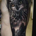 Фото тату на руке: реалистичное изображение совы, вписанной в горный пейзаж с еловым лесом, выполненное в черно-серых тонах.