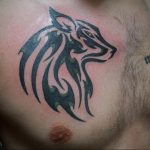 Фото тату племенного волка на груди. Черно-зеленый рисунок с геометрическими узорами и стилизованной головой волка, символизирующий силу и свободу.