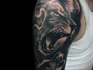 Фото тату рычащего льва на плече. Детализированный черно-серый рисунок с открытой пастью и свирепым взглядом, дополненный темными узорами.