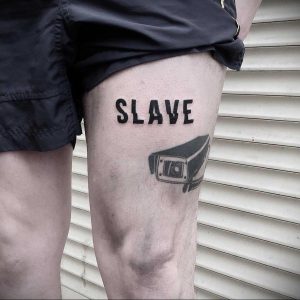 Фото татуировки на бедре с надписью "SLAVE" и изображением автомобильной фары, символизирующей зависимость и контроль. Черно-белый рисунок.