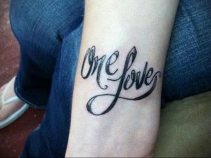 Фото татуировки с надписью 'One Love' на запястье. Черный цвет, каллиграфический шрифт, стильный дизайн.
