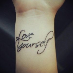 Фото татуировки с надписью 'Love Yourself' на запястье. Черный цвет, каллиграфический шрифт.