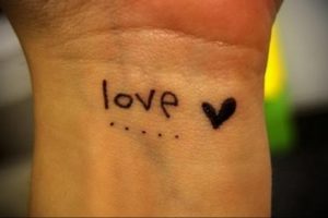 Фото простой татуировки на запястье с надписью 'love' и небольшим сердцем. Черно-серый рисунок, выполненный в наивном стиле.
