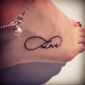 Фото минималистичной татуировки на стопе с надписью 'Love', вписанной в символ бесконечности. Черно-серый рисунок.