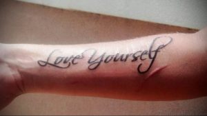 Фото татуировки на предплечье с надписью 'Love Yourself'. Черно-серый рисунок, выполненный каллиграфическим шрифтом.