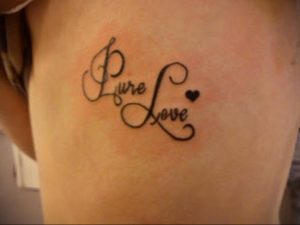 Фото тату надписи "Pure Love" выполненной каллиграфическим шрифтом с маленьким сердечком на боку девушки. Черно-белая татуировка, нежный дизайн.