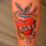 Фото тату яркого сердца с летящей птицей и надписью 'Love' на ленте. Цветная татуировка в стиле олдскул.