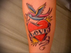 Фото тату яркого сердца с летящей птицей и надписью 'Love' на ленте. Цветная татуировка в стиле олдскул.