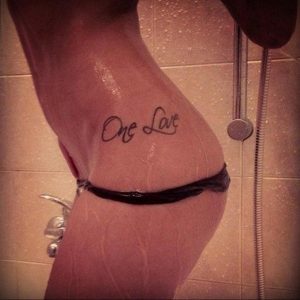 Фото тату надписи 'One Love' на боку. Черно-серая татуировка, выполненная каллиграфическим шрифтом, с акцентом на плавных линиях.