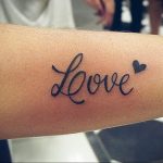 Фото тату надписи 'Love' с маленьким сердечком на руке. Черно-серая татуировка, выполненная каллиграфическим шрифтом, с акцентом на изящных линиях.