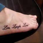 Фото тату: надпись 'Live, Laugh, Love' черными чернилами на стопе. Простой и позитивный рисунок татуировки.