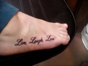 Фото тату: надпись 'Live, Laugh, Love' черными чернилами на стопе. Простой и позитивный рисунок татуировки.