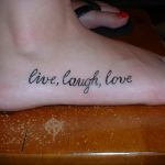 Фото тату: надпись 'Live, Laugh, Love' черными чернилами на стопе. Минималистичный и популярный рисунок татуировки.