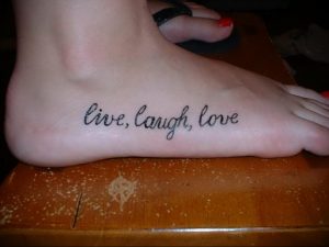 Фото тату: надпись 'Live, Laugh, Love' черными чернилами на стопе. Минималистичный и популярный рисунок татуировки.