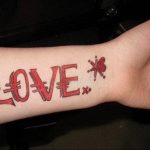 Фото тату: надпись 'Love' в ярком красном цвете, выполненная в стиле граффити с брызгами и контурами. Необычный и молодежный рисунок татуировки.