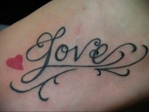 Фото тату: изящная надпись 'Love' с завитушками и маленьким красным сердцем на коже. Красивый и нежный рисунок татуировки для девушек.