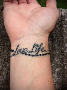 Фото тату с надписью 'love life' и небольшими цветочными элементами на запястье. Черно-желтая татуировка в каллиграфическом стиле.
