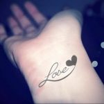 Фото татуировки надписью 'Love' и маленьким сердечком на запястье. Черно-белый рисунок татуировки с изящным шрифтом.