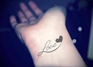 Фото татуировки надписью 'Love' и маленьким сердечком на запястье. Черно-белый рисунок татуировки с изящным шрифтом.
