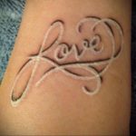 Фото белой татуировки надписью 'Love' в форме сердца на запястье. Минималистичный и изящный рисунок татуировки.
